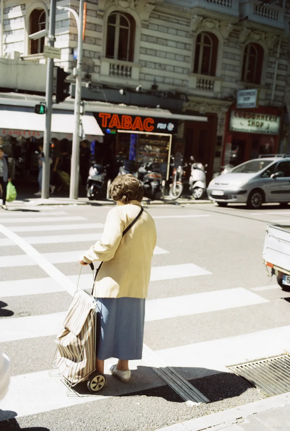 A woman with a shopping trolley waiting at a pedestrian crossing, a tabac behind her, on Kodak Vision3 50D, scanned on a Fuji Frontier.