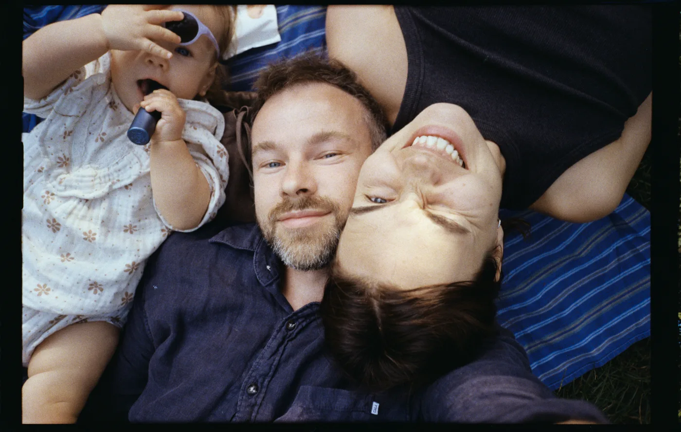 A family selfie on a striped blanket: a bearded man, a woman upside down beside him laughing, and a toddler in sunglasses.