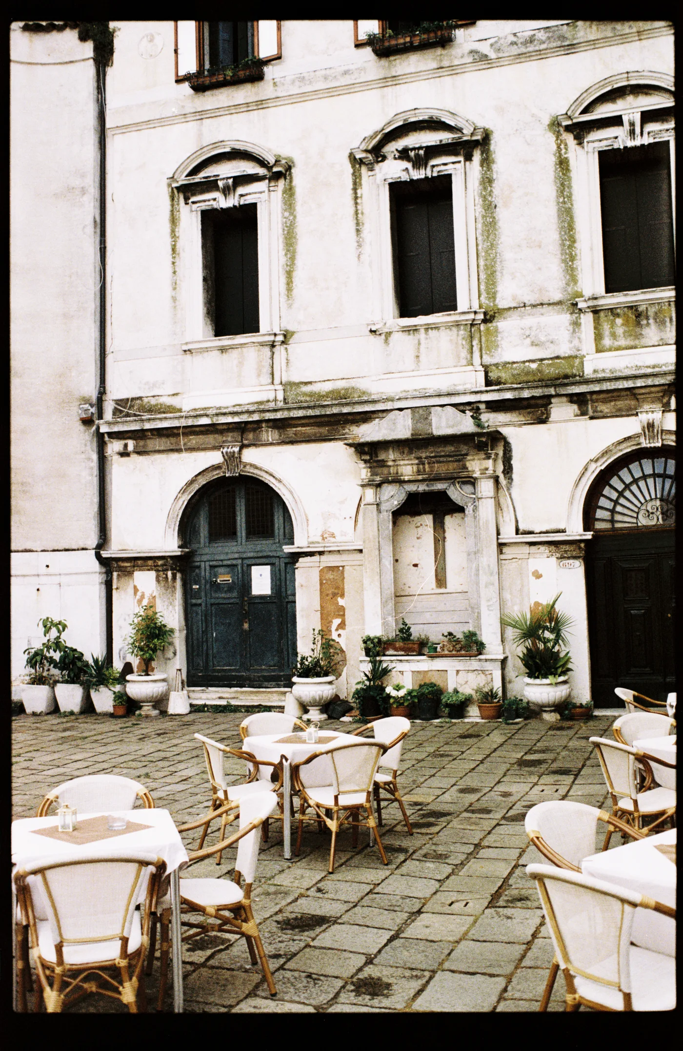 Café tables with white cane chairs on a paved square in front of a weathered palazzo with arched doors and potted plants.