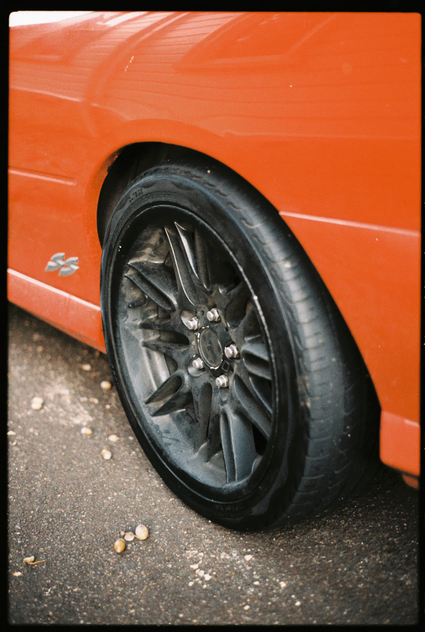 The rear wheel of an orange car, a dark alloy rim on grey asphalt scattered with acorns.