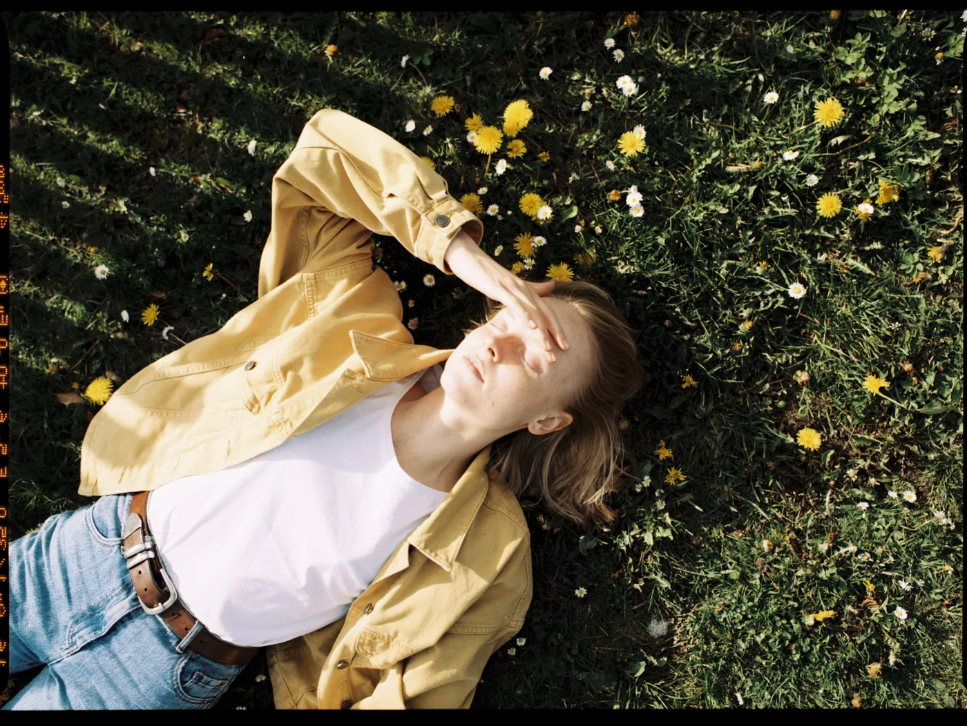 A woman lying in grass among dandelions and daisies, shading her eyes from the sun, in a mustard shirt and jeans.