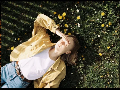 A woman lying in grass among dandelions and daisies, shading her eyes from the sun, in a mustard shirt and jeans.