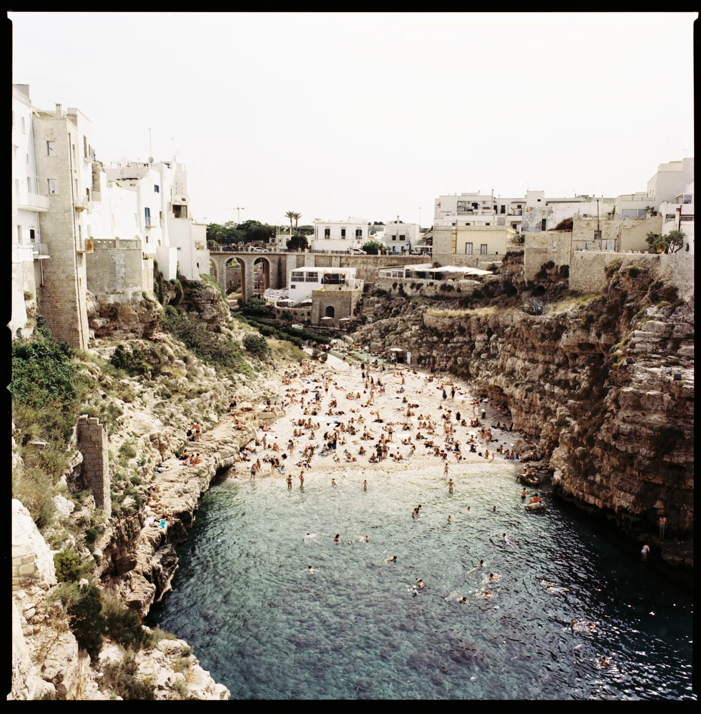 A crowded pebble beach in a narrow cove between limestone cliffs, white houses above.