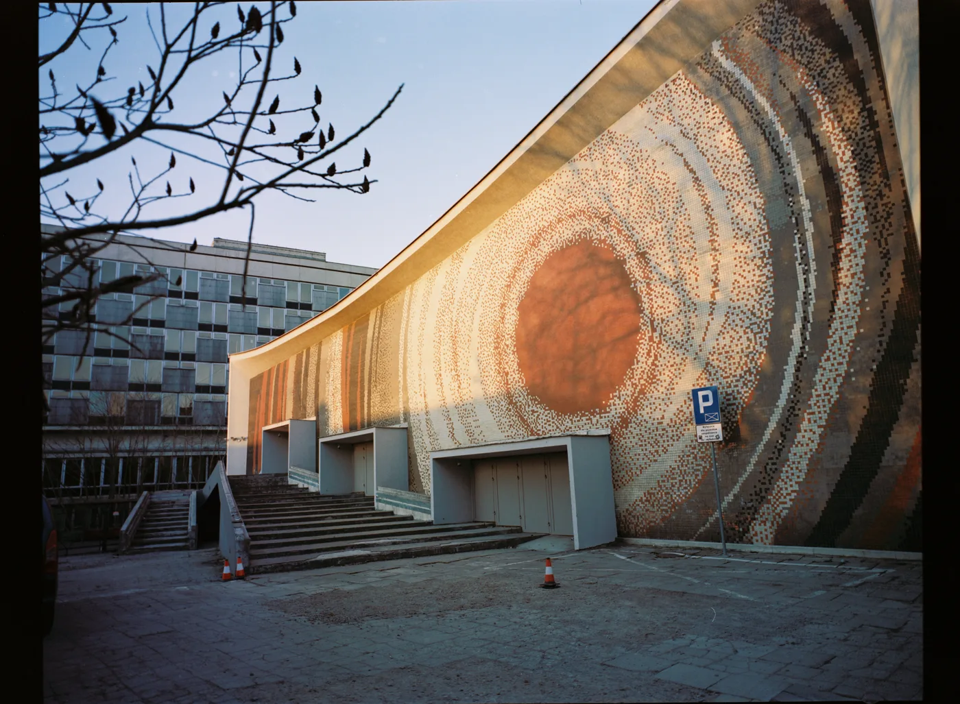 A curved modernist building faced with a mosaic of concentric rings around an orange sun, bare branches in front.