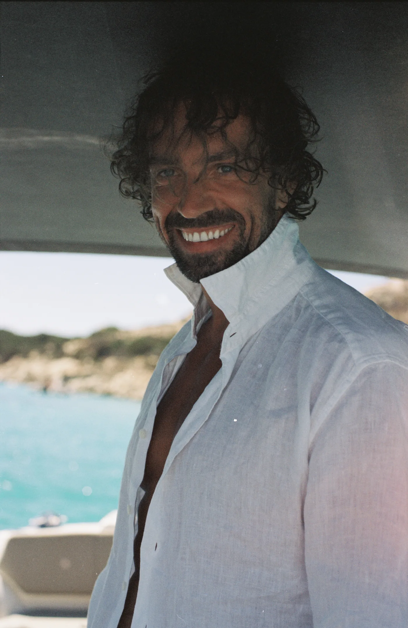 A laughing man with curly hair and an open white linen shirt under a boat's canopy, turquoise sea behind him.