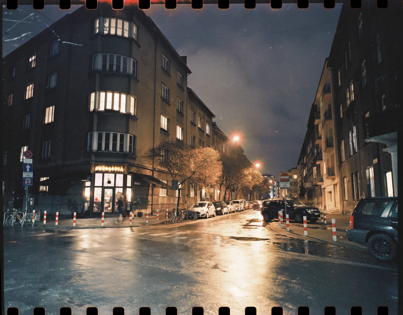 A city street corner at night, wet asphalt reflecting orange street lights between tall apartment blocks.