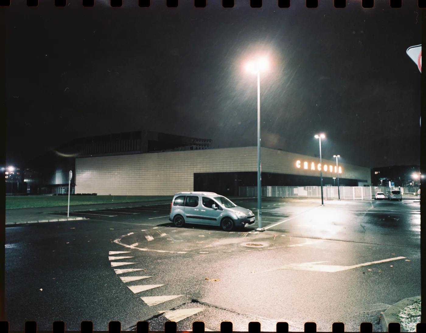 A wet car park at night under street lamps, a silver van parked before a long low building lettered CRACOVIA.